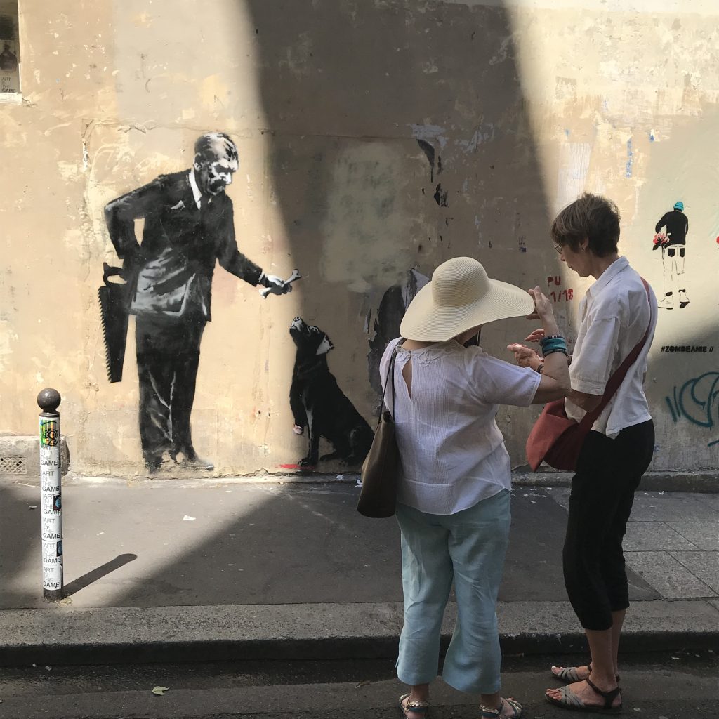 Banksy mural in Paris showing a businessman offering a bone to a three-legged dog while hiding a saw behind his back, located near the Sorbonne University.