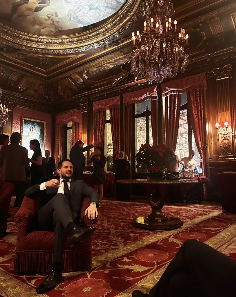 Well-dressed man seated in an armchair, drinking wine in the spacious and refined dining room of the Travellers Club in Paris.